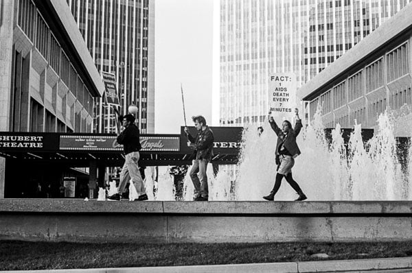 A black and white photograph from the early '90s shows protestors amid tall buildings. One holds up a sign that reads: "Fact: 1 AIDS death every 7 minutes."