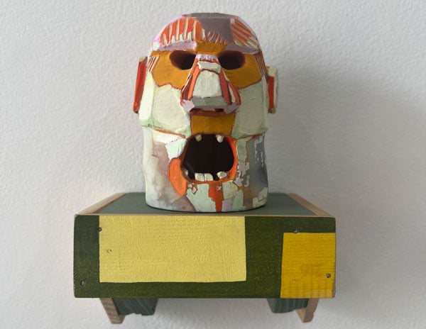 A small ceramic head perched on a shelf is made of a mix of colors and has an open mouth with stubby canines.