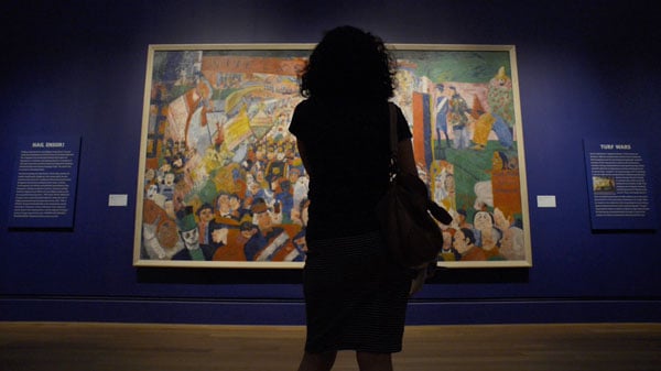 The figure of a woman is is silhouetted as she stands before a bright painting with many figures in a gallery.