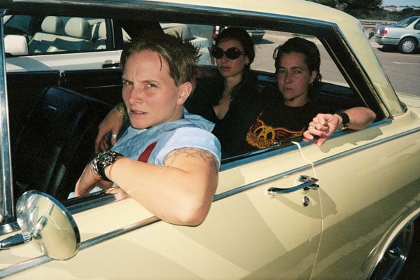 A color photograph shows a pair of butch women and femme wearing sunglasses looking cool in a yellow Chevy Nova