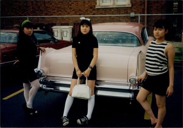 A vintage image shows three Asian women, all members of a band, leaning against a classic car painted pink.