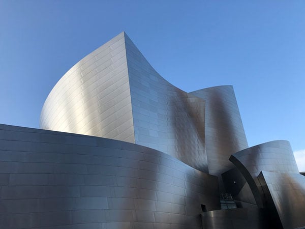 A color photograph shows an undulating building clad in stainless steel panels framed by a blue sky.