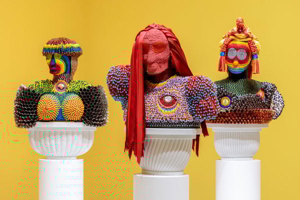 Three beaded sculptures evoking Native dress rest on white plinths before a bright yellow wall