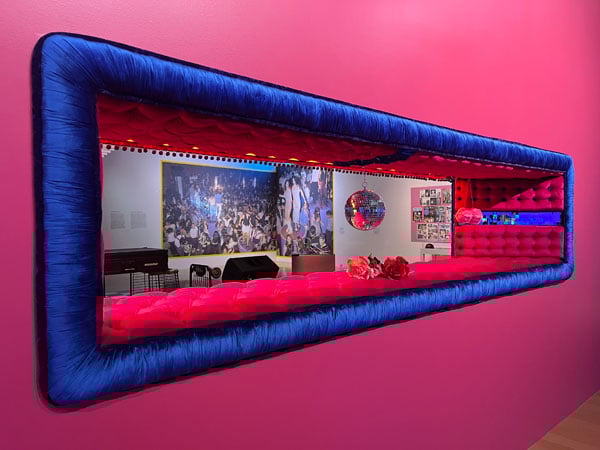 A gallery wall, painted bright pink, features a cut-out that offers views of another gallery space and is lined in bright red and blue upholstery typical of lowriders.