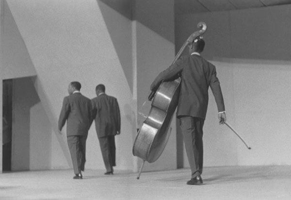 A black and white photograph by Roy DeCarava shows three Black men in suits, one carrying a string bass, walking away from the camera.