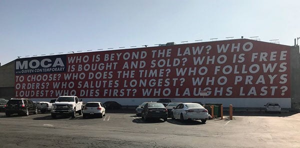 A view of Barbara Kruger's mural on the side of MOCA reads: WHO IS BEYOND THE LAW? WHO IS BOUGHT AND SOLD? WHO IS FREE TO CHOOSE? WHO DOES THE TIME? WHO FOLLOWS ORDERS? WHO SALUTES LONGEST?...