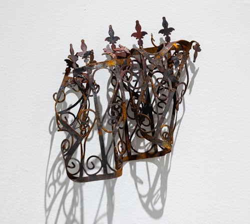 A steel sculpture by Margaret Griffith, resembling decorative ironwork, hangs on the white wall of a gallery, its forms bent and singed by fire.