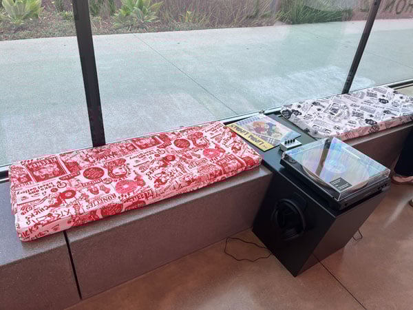A museum bench against a window features is upholstered in a fabric resembling a punk zine. Attached to it is a record player listening station.