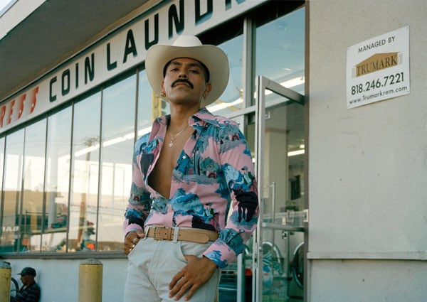 A color photograph shows a Mexican man in bright pink and blue shirt open to his waist. He wears a white cowboy hat and stand before a coin laundry.