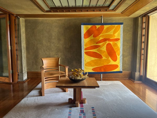 A room in Frank Lloyd Wright's Hollyhock house features a wood chair, a table bearing a bowl with dried flowers and, as backdrop, a colorful textile featuring oblong abstract forms in orange and yellow.