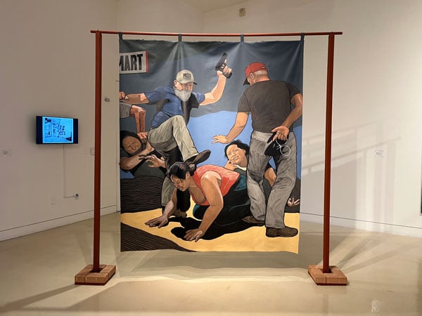 A painting hanging from a wooden frame like a curtain depicts ICE agents kicking a Mexican-looking woman who is on her hands and knees