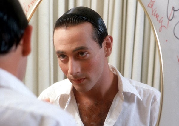 A photograph from the doc Pee Wee as Himself shows the actor Paul Reubens wearing a white shirt, his hair slicked back, looking in a mirror.