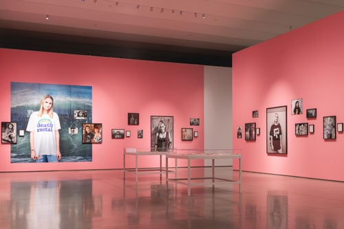 An installation view of a museum shows a gallery with bright pink walls hung with photographs of teen girls.