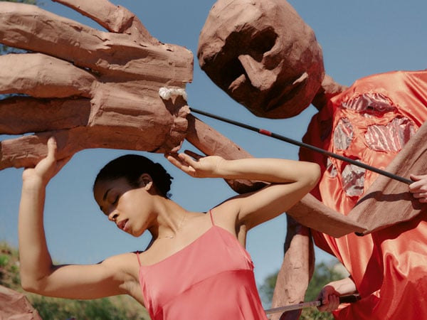 A dancer in a pink gown raises up her arms to protects herself from a giant puppet reaching forward with a massive hand.