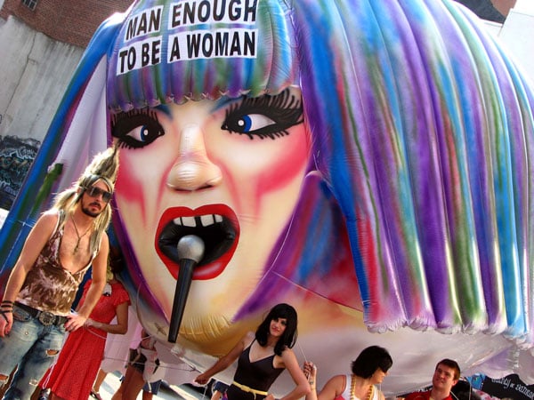 A giant inflatable carried through a city street shows the head of a gender-bendy figure with a blue/purple wig singing into a microphone. On the figure's head is the phrase: "Man Enough to Be a Woman."  Before the balloon stands a man in a low-cut tank top and spiked hair.