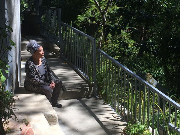 The artist Betye Saar, a diminutive Black woman in a headwrap and loose blouse, sits on a step overlooking a lush garden.