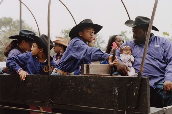 A photo by Rahim Fortune shows a Black family sitting in a wagon in matching purple-blue shirts and black cowboy hats. The child at center gazes at the camera.