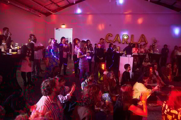 A room is filled with people at a party. On a wall behind them are inflatable balloons that spell out the word "CARLA."