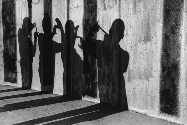 A black and white photograph by Graciela Iturbide shows the silhouettes of four figures throwing up gang signs against a wall.
