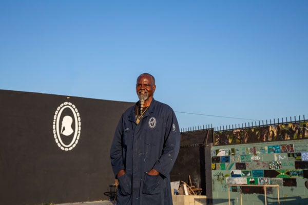 A color photograph shows a smiling Black man with a long braided beard and overalls standing outside what appears to be an artsy warehouse space.