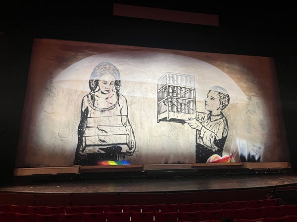 A giant paintings taking the form of a theater curtain by Enrique Martinez Celaya shows a young boy holding up a caged bird next to a pensive-looking bird also holding another birdcage.