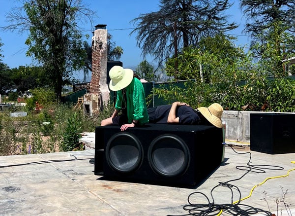 Two people wearing large straw hats that obscure their faces rest on a giant subwoofer speaker resting on the foundation of a house that burned down.