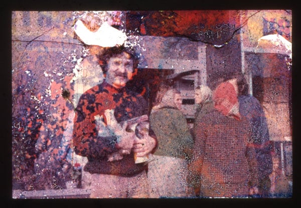 A damaged slide photograph shows an image of a mustachioed man in a sweater clutching some vases as a group of women in headscarves chat behind him.