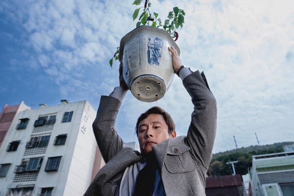 A Korean man in a jacket and tie is seen holding a potted plant over his head, as if he is going to throw it.