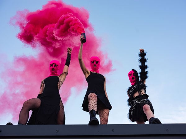 Three members of the band Pussy Riot, wearing black dresses and pink balaclavas, raise their arms in defiance — one holding a can that emits pink pigment.