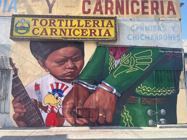 A mural shows a small child with Indigenous features standing next to an adult in a mariachi outfit who has been handcuffed.