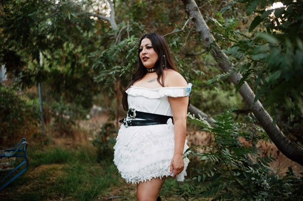 A portrait shows the singer San Cha, who is Latina, in a white dress with black leather belts, standing in a wooded area.