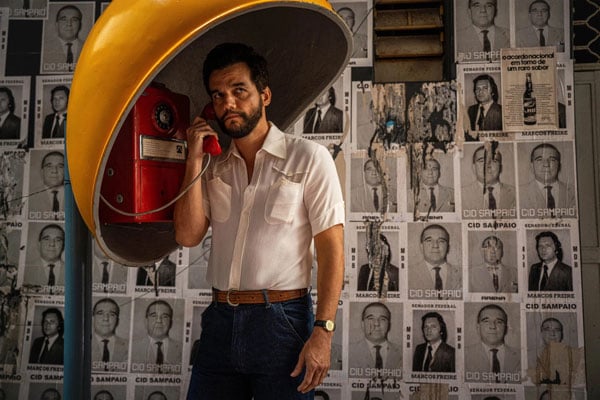 A film still shows actor Wagner Moura talking into a 1970s-style pay phone as he stands before a wall of black and white portraits of politicians.