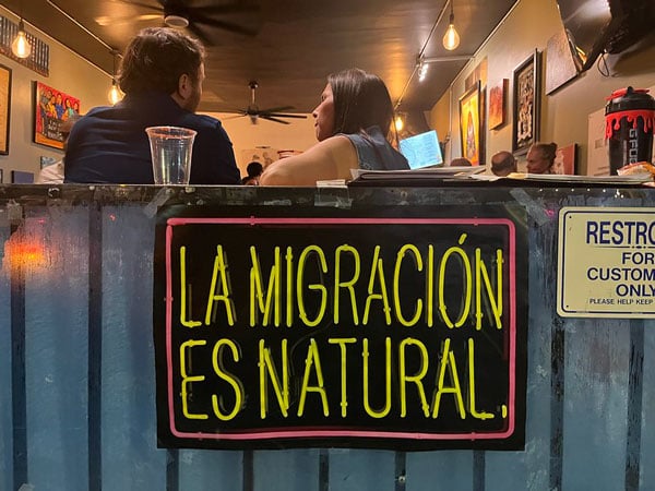 A poster by Patrick Martinez hangs on a wall outside a bar and reads "La Migración es Natural" in bright colors.