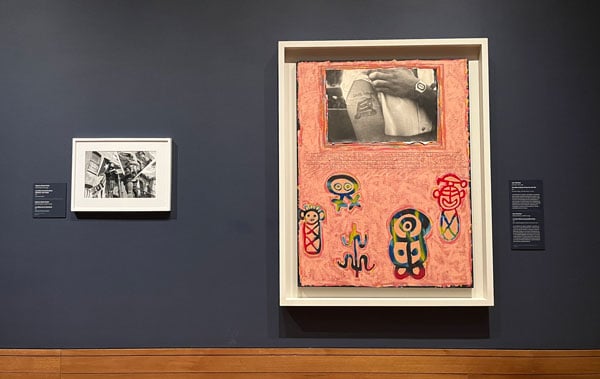 A view of a gallery wall shows a framed black and white photograph and pink painting that features a black and white photograph of a man lifting his sleeve at top. The painting also contains petroglyphyic signs from Puerto Rico.
