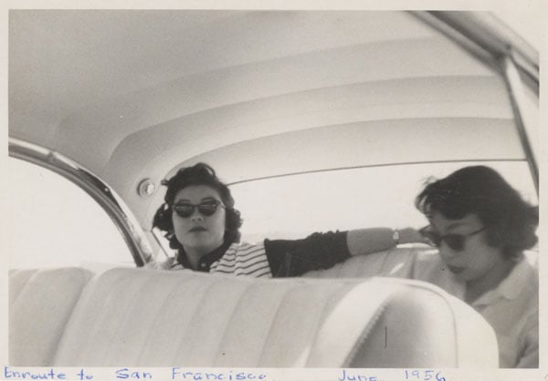 A vintage black and white snapshot from the 1950s shows two Japanese American women in cat eye sunglasses sitting in the backseat of a Bel Air.