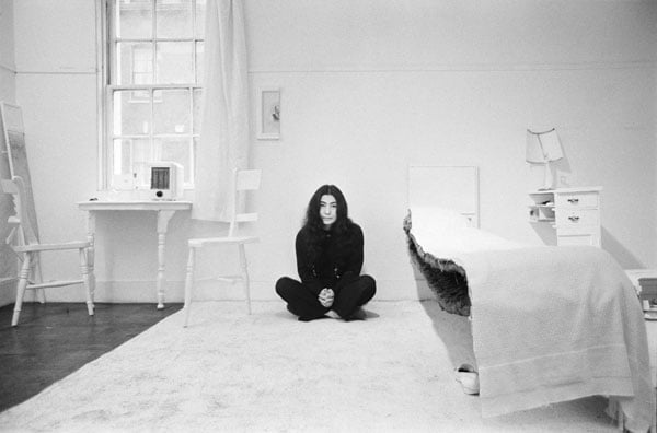 A black and white photo shows Yoko Ono, dressed all in black, in a room with white objects cut in half — like a bed, a chair, a table and a nighttable.