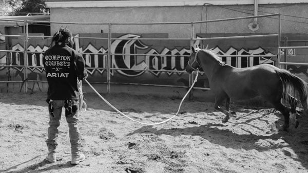 A Black man with locs wearing a jacket that reads "Compton Cowboys x Ariat" trains a horse in a corral