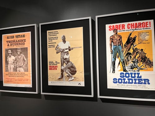 A row of Blaxploitation movie posters on a darkened gallery wall feature flyers for Western themed films like "Thomasine and Bushrod" and "Soul Soldier"
