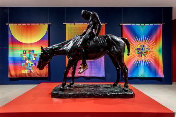 A vintage turn of the 20th bronze sculpture showing a slumped Indigenous man on a horse sits on a red plinth before three flags in psychedelic colors.