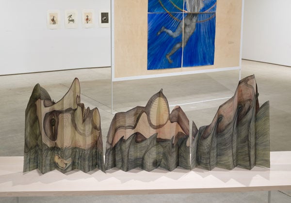 A piece of paper, folded accordion style, reveals a drawing of a reclining woman, her profile and body resembling a mountain range.