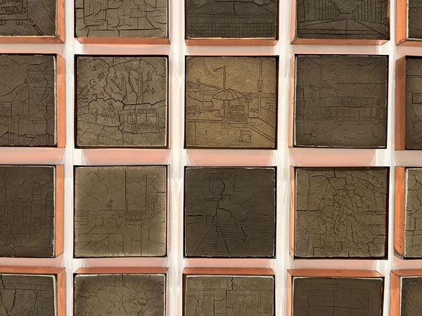 A wall installation by Jackie Amezquita shows rows of square panels crafted from soil with drawings incised on the hardened surface.