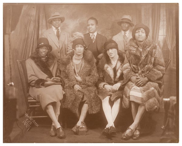 A vintage portrait from the 1920s shows three Black drag kings seated behind four drag queens in a portrait studio.