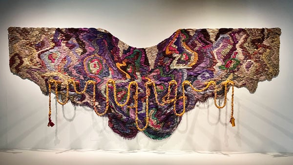 A textile presented on a wall is composed of rich shades of purples and brown and bears a series of extended arms holding a bright yellow piece of knotted fabric.