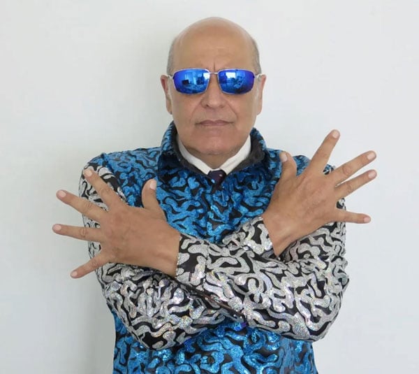 The Dark Bob, an LA performance artist poses in a sequin jacket with a pattern of silver and blue and wears blue sunglasses.