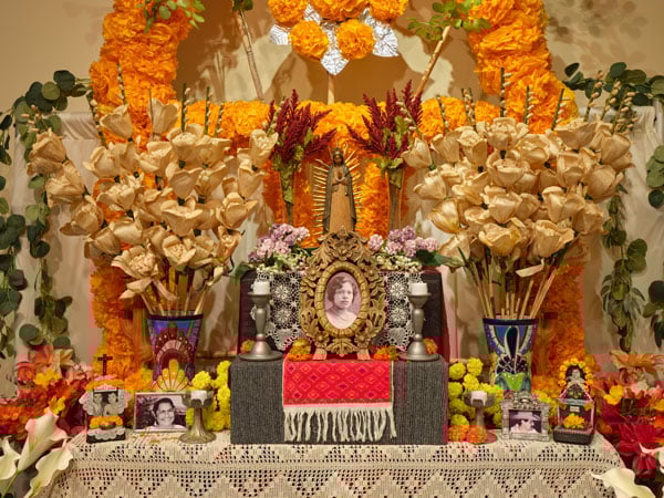 A close up of an altar for Day of the Dead shows family photos in frames surrounded by colorful vases fillowed with flowers, framed by orange flowers made with paper.
