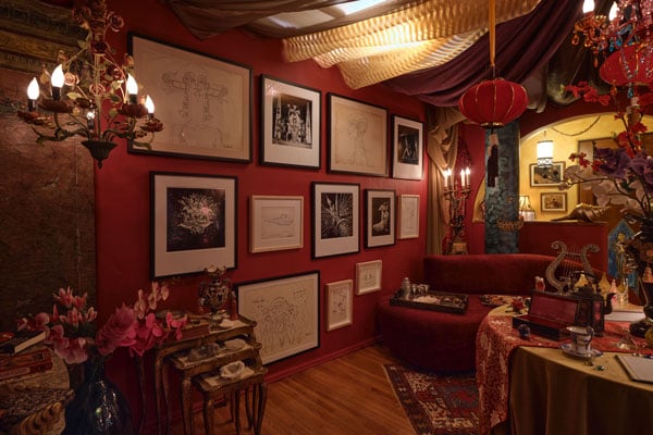 An ornate room has walls painted a scarlet red along with a sofa of the same color. Hanging salon-style on a wall are black and white photos and a series of drawings.