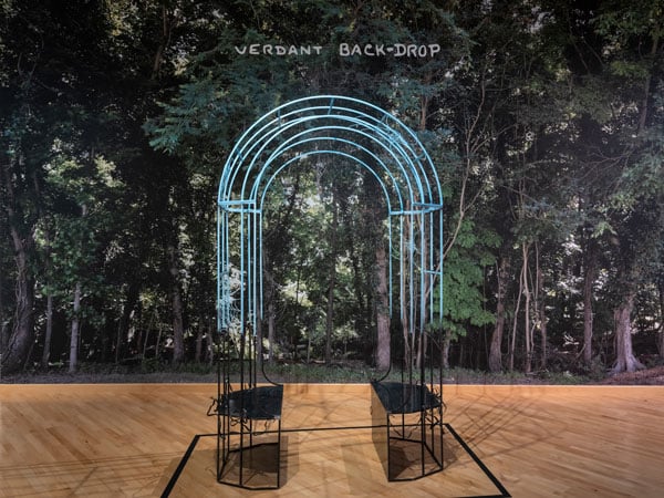 A gazebo-style arch illuminuted with neon stands before a leafy green image of a forest, over which is written the phrase "Verdant Back-Drop."