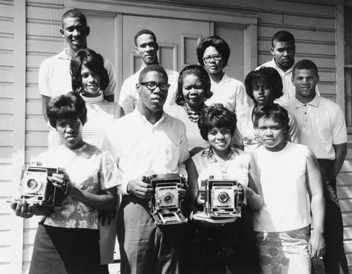 A vintage black and white photo from 1961 shows a dozen Black people standing in rows, several of them holding cameras.