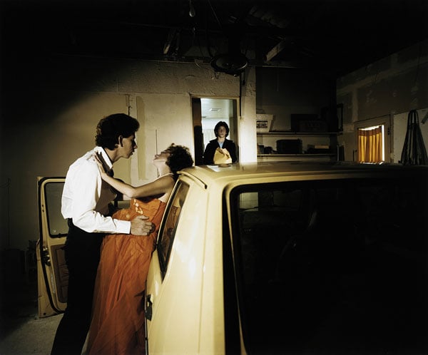 A staged photograph created by Eileen Cowin shows a man looking as if he is about to kiss a woman who leans on a car. In the distance, another woman comes through the doorway.