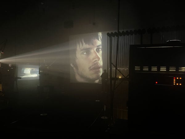 A darkened room shows various projection mahcinery in the foreground. At center, a beam of light projects Gael García Bernal's face on a wall.
